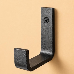 Puede incluir: Un gancho de metal negro en forma de J con una superficie texturizada. El gancho está montado con un solo tornillo. El diseño es simple e industrial, adecuado para colgar objetos. El gancho está sobre un fondo beige claro.