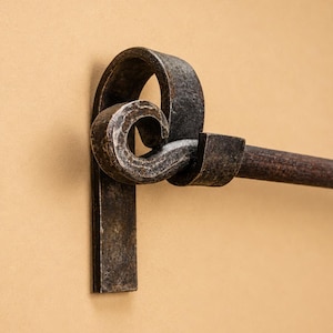 May include: A dark, textured metal towel rail featuring a hand-forged bracket with a decorative scroll design. The mounting plate is flat and rectangular, connecting to a curved, looped metal piece that holds a horizontal rod with a dark finish.