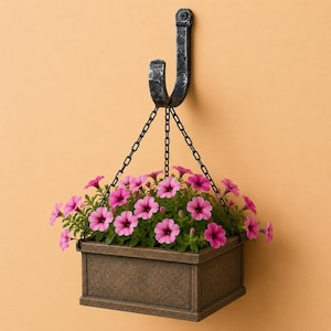 May include: A hanging planter filled with bright pink flowers. The rectangular planter is made of a textured, brown material and is suspended by a chain from a decorative, dark metal hook attached to the wall.