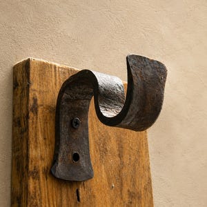 Puede incluir: Un gancho de metal oscuro y desgastado con un diseño curvo en forma de U, montado sobre una pieza rectangular de madera de color marrón claro con textura. La superficie metálica muestra signos de oxidación y una sola tornillo negro.