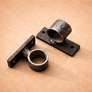 May include: Two dark metal curtain rod holders. One holder is a ring attached to a rectangular plate with two holes. The other holder is a cylinder attached to a rectangular plate with two holes. The holders are on a tan surface.