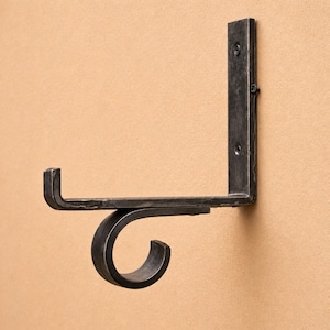 Op de afbeelding: Een donkergrijze, L-vormige metalen plankbeugel. De beugel heeft een platte horizontale plank en een verticaal steundeel, beide bevestigd aan een muur. De beugel heeft een decoratief, gebogen haakontwerp. Schroeven bevestigen de beugel aan de muur.