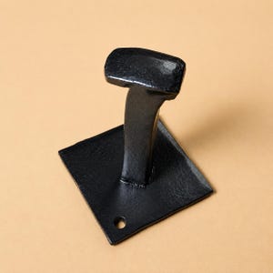 May include: A black, forged metal hook with a square base and a curved stem leading to a rectangular top. The base has a single hole. The hook is set against a beige background.
