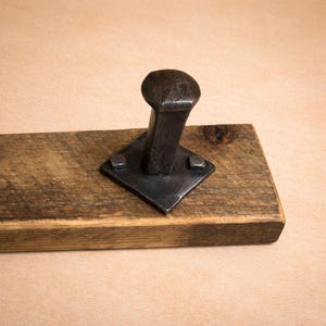 Puede incluir: Un perchero artesanal y rústico. El gancho es de metal forjado oscuro y está montado sobre una pieza rectangular de madera reciclada. El gancho de metal está fijado a una placa cuadrada de metal con remaches. La madera tiene un aspecto natural y envejecido.