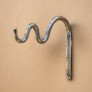 Puede incluir: Un gancho de metal para pared con un diseño ondulado y serpenteante con un acabado plateado desgastado. El gancho cuenta con una placa de montaje vertical plana asegurada con dos tornillos visibles y un brazo curvo para colgar objetos.