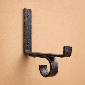 Puede incluir: Un gancho de pared de hierro forjado con un acabado oscuro y texturizado. El gancho está montado en la pared y presenta un diseño curvo para colgar objetos. Se fija a la pared con dos tornillos.