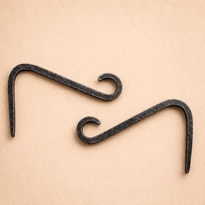 Puede incluir: Dos ganchos para plantas de hierro forjado con un acabado oscuro y texturizado. Cada gancho tiene un diseño curvo en forma de voluta en un extremo y un gancho que mira hacia abajo en el otro. Los ganchos están diseñados para colgar plantas u otros objetos decorativos.