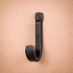 May include: A black, hand-forged iron hook. The hook is J-shaped with a curved end and a flat top secured with a screw. The hook is mounted on a neutral-toned background.