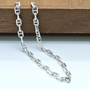 Op de afbeelding: Een zilverkleurige ketting met een reeks in elkaar grijpende schakels. De ketting wordt getoond tegen een houten oppervlak en een witte achtergrond. De schakels hebben een kenmerkende vorm, waardoor een stijlvol accessoire ontstaat.