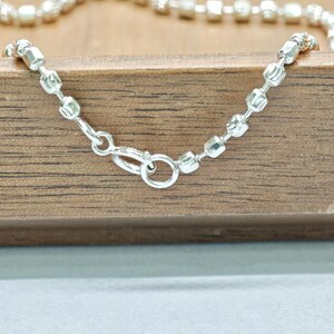 May include: A silver necklace with a delicate chain of small, square beads. The necklace is fastened with a lobster clasp. The jewellery is presented on a wooden surface.
