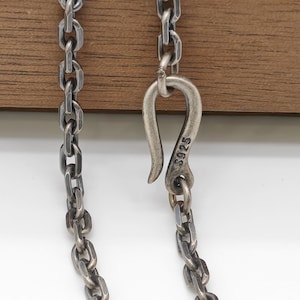May include: Close-up of a silver chain necklace with a hook clasp. The chain links are rectangular and connected. The hook clasp is marked with "S925". The background is a wooden surface.