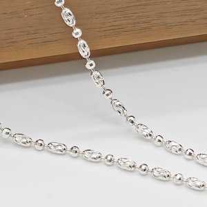 May include: A silver necklace with a delicate chain design. The necklace features a pattern of small, round beads alternating with elongated, textured oval beads. The chain is displayed against a white surface and a wooden background.