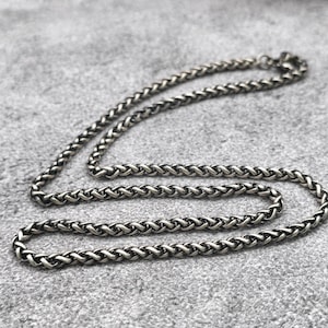 May include: A silver-colored chain necklace with a woven design. The necklace is lying on a gray surface. The chain is made of small, interlocking links, creating a textured appearance. The necklace is a jewelry item.
