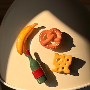 Könnte beinhalten: Eine Sammlung von Miniatur-Lebensmitteln, darunter eine Banane, eine Brezel, eine Weinflasche und ein Käsewürfel. Die Banane ist gelb-orange, die Brezel ist braun mit weißen Punkten, die Weinflasche ist grün mit rotem Etikett und der Käse ist orange.