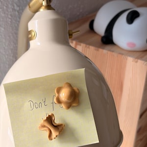 Könnte beinhalten: Cremefarbene Schreibtischlampe mit einem verstellbaren Arm und Akzenten in Messingfarbe. Ein gelber Haftzettel ist an der Lampe befestigt mit dem handgeschriebenen Text "Don't f..." und zwei dekorativen Magneten. Eine Panda-Figur liegt auf einem Holzregal.