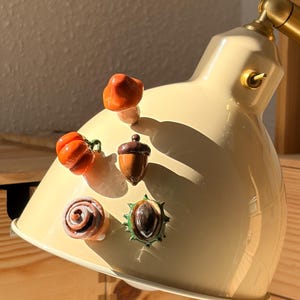 Puede incluir: Una pantalla de lámpara de metal color crema decorada con cuatro pequeños adornos de cerámica brillante: un hongo naranja, una calabaza naranja brillante, una bellota marrón y una castaña marrón dentro de una cáscara verde espinosa.