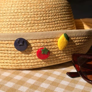 Könnte beinhalten: Ein Sonnenhut aus Stroh mit einem beigen Band, verziert mit fruchtförmigen Verzierungen: einer Blaubeere, einer Tomate und einer Zitrone. Eine Sonnenbrille ist teilweise sichtbar. Der Hut liegt auf einer karierten Oberfläche.