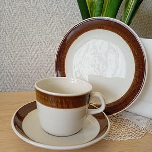 Może przedstawiać: Zestaw ceramicznej filiżanki do kawy ze spodkiem w kolorze kremowym, z szerokim, ciemnobrązowym pasem na krawędzi filiżanki i spodka. Brązowe pasy ozdobione są delikatnym, pionowo prążkowanym wzorem o wyczuwalnej fakturze.