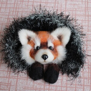 May include: A handmade felt red panda brooch. The red panda has a white face with black eyes and a black nose. The brooch is surrounded by a black and silver fringe.