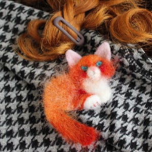 Op de afbeelding: Een handgemaakte oranje en witte vilten kattenbroche met blauwe ogen, liggend op een zwart-wit pied-de-poule stof. Een grijze haarclip is zichtbaar op de achtergrond. Een charmant accessoire.