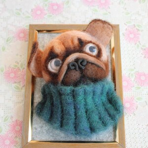 Può includere: Un'opera d'arte in feltro incorniciata raffigurante un carlino con un viso marrone e marrone scuro, grandi occhi e un naso scuro. Il carlino indossa un maglione verde acqua e blu. L'opera d'arte è in una cornice di legno.