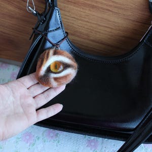 Op de afbeelding: Een ronde, naaldgevilte hanger met een realistisch geel glazen oog met een verticale zwarte pupil, omringd door bruine, witte en beige wolvezels. De hanger hangt aan een dun bruin koord en wordt getoond tegen een zwarte handtas.