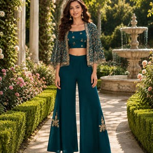 Könnte beinhalten: Ein dreiteiliges Outfit in Petrol, bestehend aus einem ärmellosen bauchfreien Oberteil, einer weiten Hose und einem passenden Umhang. Oberteil und Umhang sind mit goldener Blumenstickerei verziert, die Hose hat goldene Motive am Saum.