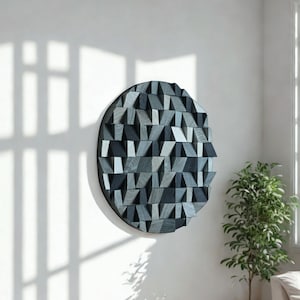 May include: A round, geometric wall art piece with a dark gray frame. The artwork features a textured surface composed of various-sized, angled wooden blocks in shades of gray and blue. The piece is mounted on a white wall, with sunlight casting shadows.