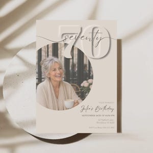 Op de afbeelding: Een beige verjaardagsuitnodiging met een groot reliëfcijfer 70 en het woord seventy in cursief schrift. Een ronde foto toont een glimlachend persoon met een kopje. De tekst bevat You're invited to Julie's Birthday, 16 september om 20:00 uur.