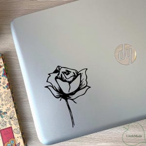 Op de afbeelding: Een zwarte rozensticker is bevestigd op een zilveren laptop. De sticker is een eenvoudige lijntekening van een roos met een steel. De laptop heeft een HP-logo in de rechterbovenhoek.