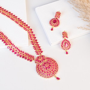 May include: A gold-toned necklace and earring set featuring ruby-colored gemstones. The necklace has a large, circular pendant and a delicate chain. The earrings are teardrop-shaped and feature matching gemstones. The jewelry is displayed on a white surface.