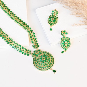 May include: A gold-toned necklace and earring set featuring emerald-green gemstones. The necklace has a large, circular pendant and a delicate chain. The earrings are teardrop-shaped. The jewelry is displayed on a white surface.