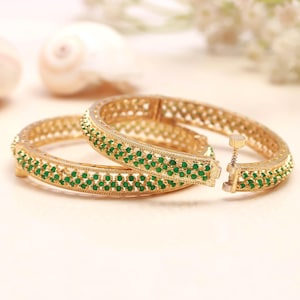 May include: Two gold-toned hinged bangles featuring a central band of small, round, vibrant green stones. The metalwork displays a delicate, openwork lattice pattern along the edges, and each bracelet includes a small, cylindrical screw-style clasp.