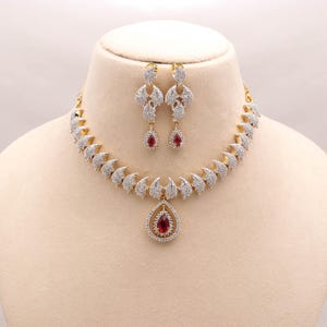 Puede incluir: Un conjunto de collar y pendientes en tono dorado con piedras transparentes en pavé dispuestas en forma de hoja. El collar incluye un colgante central en forma de lágrima con una piedra roja, a juego con los dos pendientes colgantes.