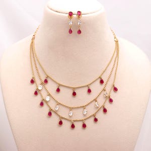 May include: A gold-toned multi-strand necklace featuring three delicate chains adorned with alternating teardrop-shaped red and clear white stones. The set includes a pair of matching drop earrings, each featuring a single red and white stone.