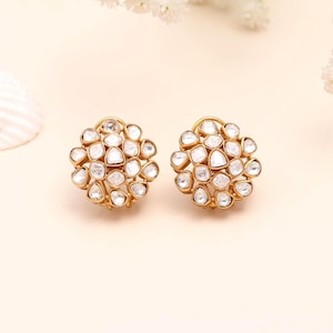 May include: A pair of gold-toned stud earrings. Each earring features a cluster of clear, irregularly shaped stones. The earrings are set against a soft, cream-colored background, with subtle floral accents in the upper right corner. The earrings are designed to be worn on the ear.