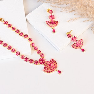 May include: A gold-toned jewelry set featuring a necklace and earrings. The necklace and earrings are adorned with vibrant pink gemstones arranged in a floral pattern. The earrings have a fan-shaped design with a teardrop-shaped gemstone pendant.