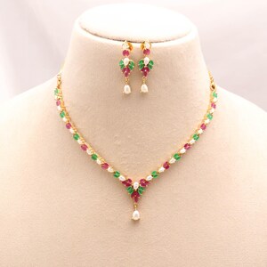 Puede incluir: Un conjunto de collar y pendientes colgantes en tono dorado con piedras rojas y verdes de corte marquesa dispuestas en un patrón floral, acentuadas con pequeñas perlas blancas. El collar tiene un colgante central y un cierre seguro.