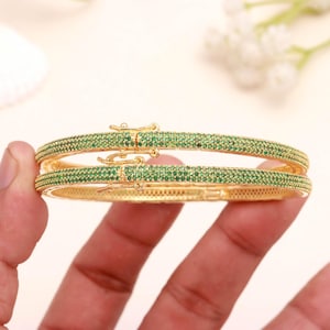 May include: A pair of gold-tone hinged bangle bracelets featuring a pavé setting of small, round, vibrant green stones covering the entire outer surface. Each bracelet includes a secure, integrated box clasp mechanism in matching gold-tone metal.