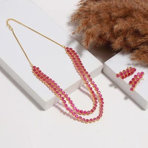 Könnte beinhalten: Ein goldfarbenes Halsketten- und Ohrringset mit mehreren Reihen leuchtender, facettierter rosa Steine. Die Halskette ist mit einer doppelsträngigen Kette gestaltet, und die Ohrringe bestehen aus einer linearen Anordnung derselben rosa Steine.