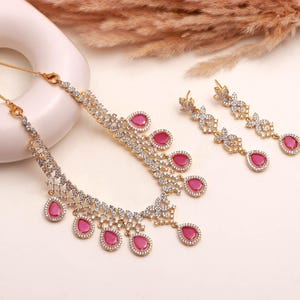 May include: A gold-toned jewelry set featuring a necklace and matching earrings. The necklace and earrings have a floral design with clear and pink gemstones. The necklace has a series of pink teardrop-shaped stones.