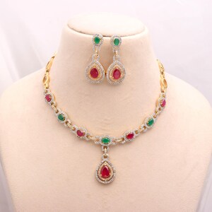 May include: A gold necklace and earring set. The necklace has alternating red and green gemstones, with a teardrop pendant. The earrings echo the pendant design, with red teardrop gems and green accents. The set is displayed on a mannequin.
