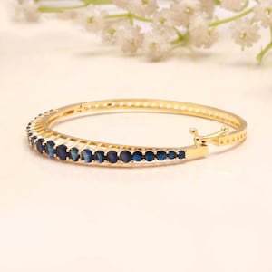 Puede incluir: Una pulsera rígida articulada de tono dorado que presenta una hilera de piedras redondas de color azul intenso engastadas a lo largo de la mitad frontal. La banda metálica tiene un acabado pulido e incluye un cierre seguro en el lateral.