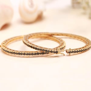 May include: A pair of gold hoop earrings featuring a row of small, round, black stones set along the outer edge. The earrings have a hinged clasp mechanism for closure, showcasing a metallic gold finish and a circular design with black accents.