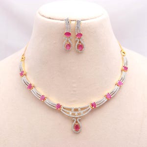 Puede incluir: Un conjunto de collar y pendientes colgantes en tono dorado con piedras rosas ovaladas y cristales transparentes en pavé. El collar presenta un diseño curvo de varias capas con un colgante central en forma de lágrima, a juego con los pendientes.