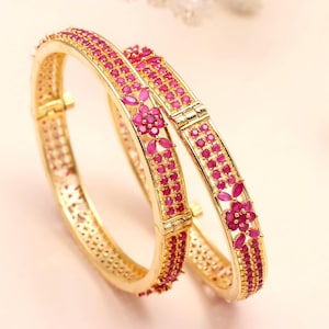 May include: Two gold-colored bangles with intricate floral and geometric designs. The bangles are adorned with numerous small, bright pink gemstones. The floral patterns are a prominent feature, adding a touch of elegance to the jewelry.