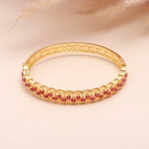 K&ouml;nnte beinhalten: Ein goldfarbenes Armreif-Armband mit einer Reihe kleiner, roter Edelsteine. Das Armband hat ein dekoratives, durchbrochenes Design auf der Oberseite. Die Edelsteine sind in einer durchgehenden Linie um das Armband angeordnet.