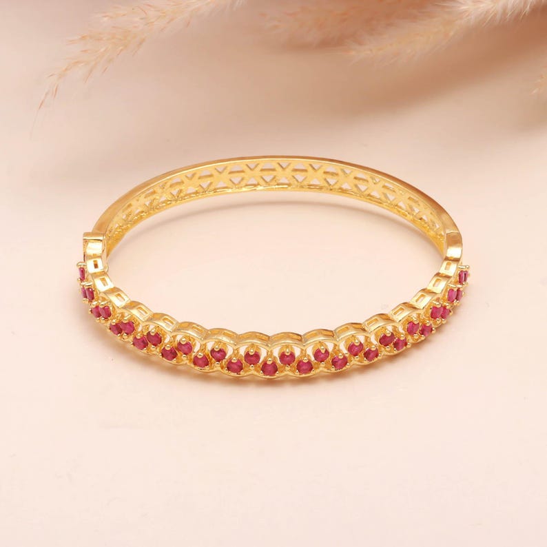 K&ouml;nnte beinhalten: Ein goldfarbenes Armreif-Armband mit einer Reihe kleiner, roter Edelsteine. Das Armband hat ein dekoratives, durchbrochenes Design auf der Oberseite. Die Edelsteine sind in einer durchgehenden Linie um das Armband angeordnet.