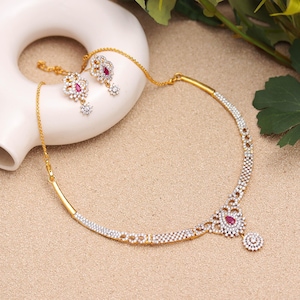 Puede incluir: Un conjunto de collar y pendientes dorados. El collar presenta un colgante central en forma de lágrima con una piedra roja, rodeado de pequeñas piedras transparentes. Los pendientes tienen un diseño similar. Las joyas se exhiben sobre una superficie neutra.