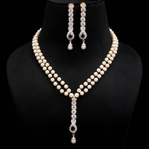 Può includere: Una collana a doppio filo di perle color crema con un pendente centrale verticale in cristalli trasparenti e una pietra nera a goccia. Gli orecchini pendenti abbinati presentano un design verticale in cristallo con una pietra nera.